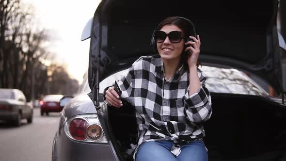 Smiling Brunette Listen to the Music in Headphone While Sitting in the Open Car's Trunk Among the