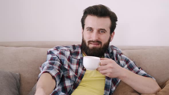 Cheerful Bearded Man with Cup Og Coffee Watching TV at Home