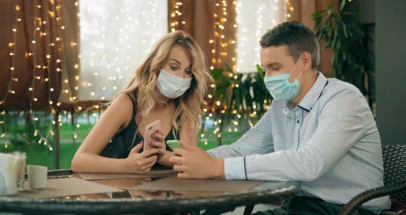 Man in Protective Mask Shows Smartphone To Woman in Cafe