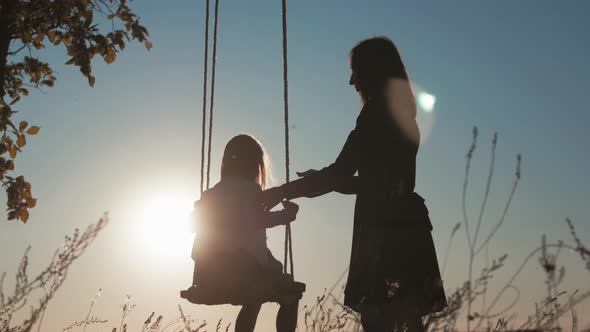 Silhouette of Happy Young Mother and Little Daughter on a Swing at Sun Light. Pretty Girl Sitting on