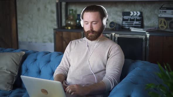 Young Bearded Man is Using Laptop and Listening to Music on Sofa at Apartment Spbd