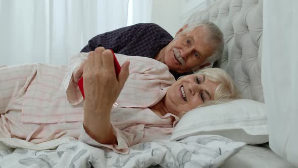 Senior Grandparents Couple Lying in Bed. Woman Getting Nervous About Man Spying Into Mobile Phone