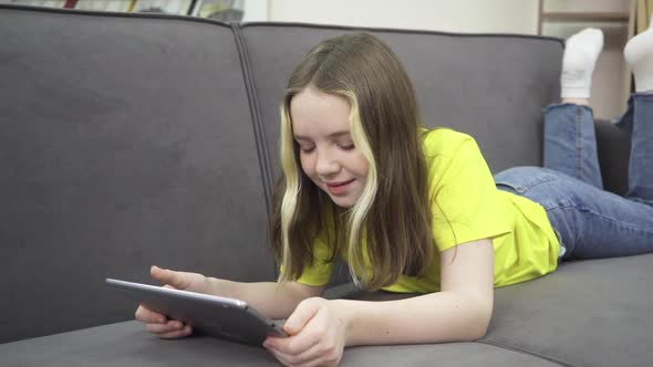 A Little Girl is Smiling While Lying on the Couch with a Tablet