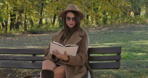 Intelligent Caucasian Woman in Sunglasses and Brown Hat Sitting on Bench and Reading the Book