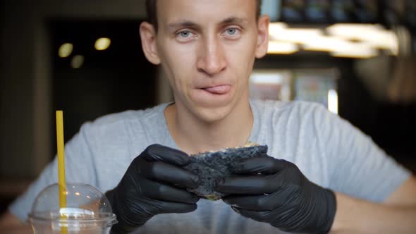 A Young Man Sits in a Restaurant and Looks Carefully at the Burger Who Holds in His Hands