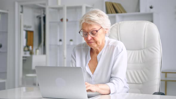 Tired sleepy old lady manager types on grey laptop