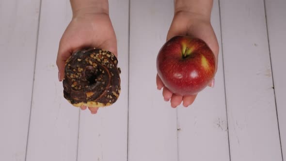 Woman Holding a Chocolate Doughnut and a Red Apple on a Light Wooden Background