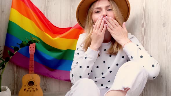 Young Blonde Millennial Hippie Woman Sending Blowing Kiss with Rainbow LGBTQ Flag at Home Balcony