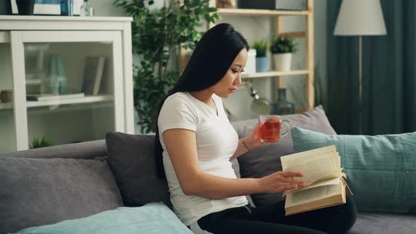 Asian Young Lady Is Reading Book and Holding Cup of Tea Then Turning Page and Drinking Sitting on