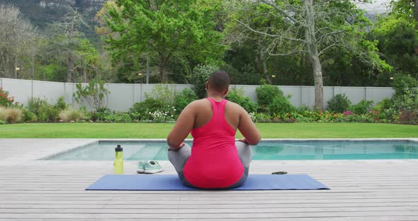 Happy african american plus size woman practicing yoga, stretching next to swimming pool in garden