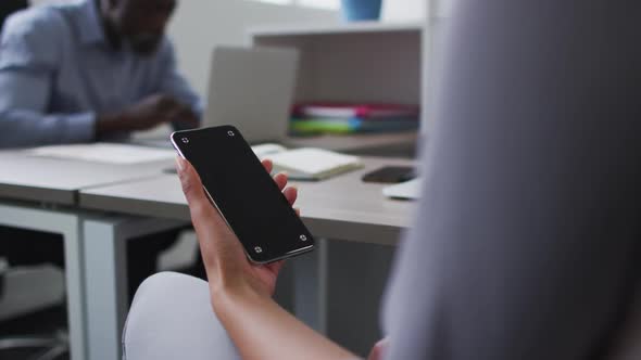 Mixed race businesswoman sitting at desk and using smartphone with copy space on screen in office