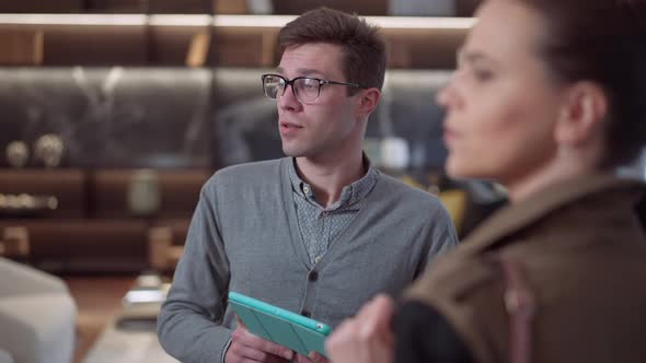 Portrait of Confident Young Male Seller in Eyeglasses with Tablet Standing in Furniture Shop Talking