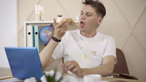 Funny Caucasian Young Man Chewing Pizza and Typing on Laptop Keyboard