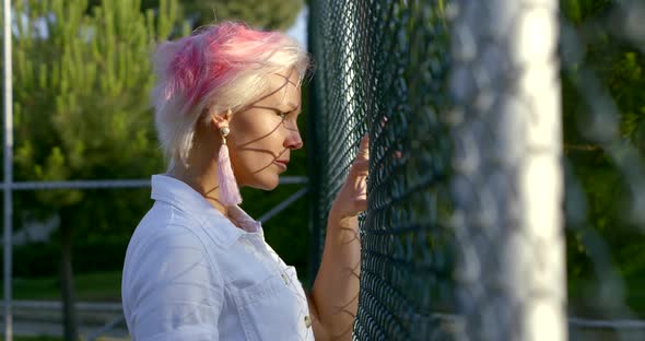 Bold and Blonde Woman Is Looking Through Metal Fence Grid of Open Sporty Area