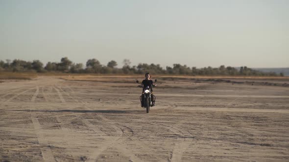 Beautiful Young Woman Riding an Old Cafe Racer Motorcycle on Desert at Sunset or Sunrise