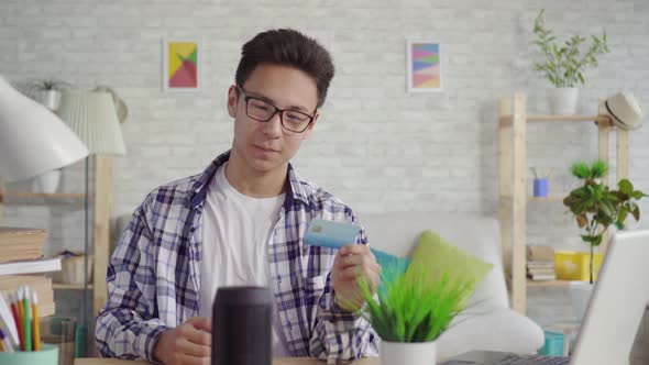 Young Asian Man in a Shirt with a Bank Card in Hand Uses a Voice Assistant