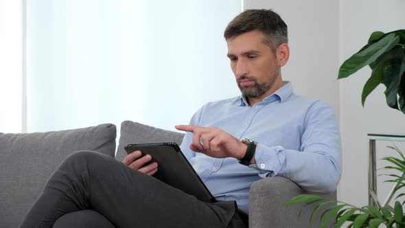 Businessman in Light Blue Shirt Sitting on the Couch Using Tablet Taping Screen