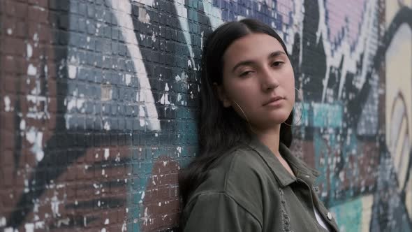 Young Beautiful Brunette Girl Stands Near the Wall with Graffiti Looking at the Camera and Smiling