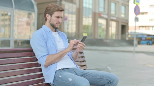 Man Reacting to Loss on Smartphone While Sitting Outdoor on Bench