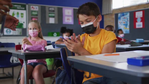 Mixed race schoolboy wearing face mask disinfecting hands in classroom, colleagues in background