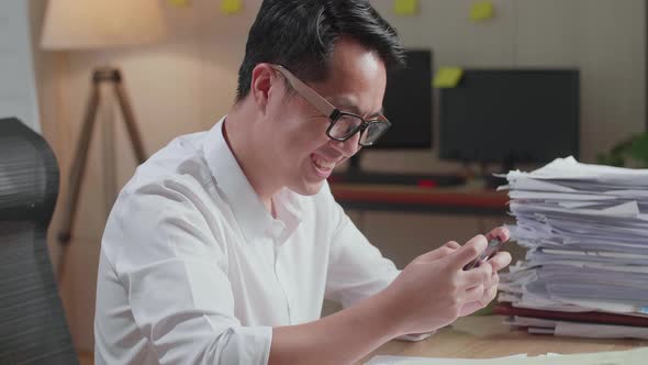 Close Up Of Asian Man Enjoys Playing Game On Smartphone After Working With Documents At The Office