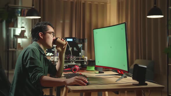 Asian Man Drinking Coffee While Work On 3D Printer And Computer With Mock Up Green Screen Display