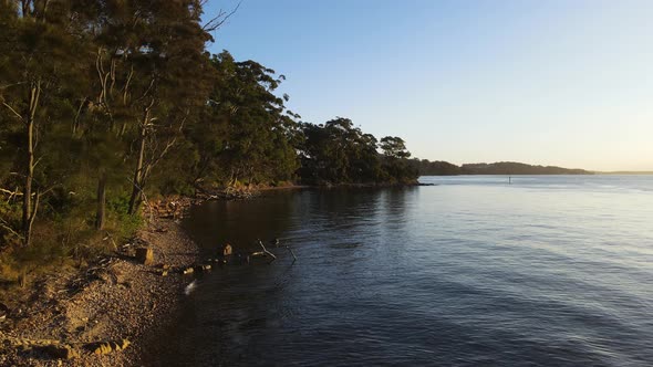 Scenic drone view travelling over calm water next to a rugged coastline covered in native bushland