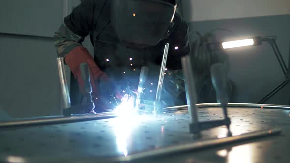 Welder in a welding helmet is welding parts in the factory in super slow motion, sparks are flying.