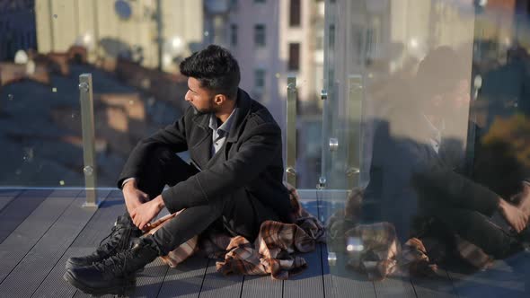 Wide Shot Anxious Troubled Middle Eastern Man Sitting at Glass Fence on Rooftop Looking Away