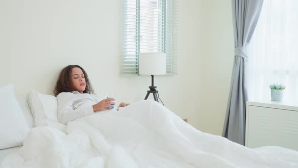 Unhappy sick Latino girl in pajamas take medicine before sleep on bed in bedroom at home.