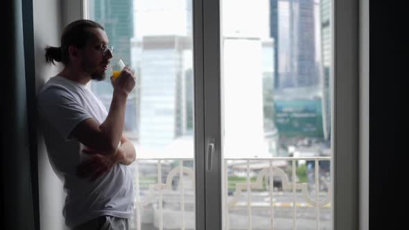 Young Guy with a Beard and Long Hair Stands at the Window and Drinks Orange Juice From a Glass