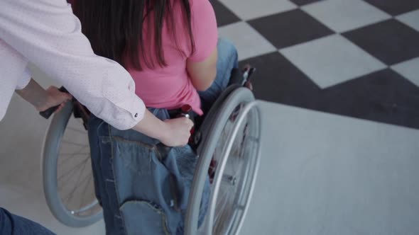 Female Helper Riding Disabled Woman Sitting on Wheelchair in Hospital Interior.