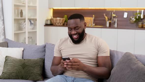 Handsome Positive African American Man in Casual Wear Sits at Home on Sofa Using His Smartphone