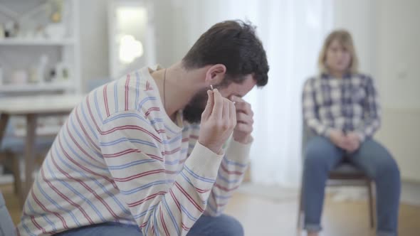 Side View of Depressed Young Caucasian Man Holding Wedding Ring and Rubbing Sore Eyes with Hand