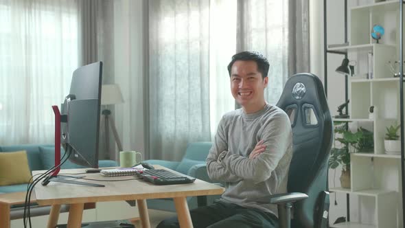 Asian Man Crossing His Arms And Smiling To Camera While Working With Desktop Computer At Home