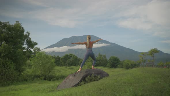Woman in warrior yoga pose on volcanic boulder with Mount Agung in background