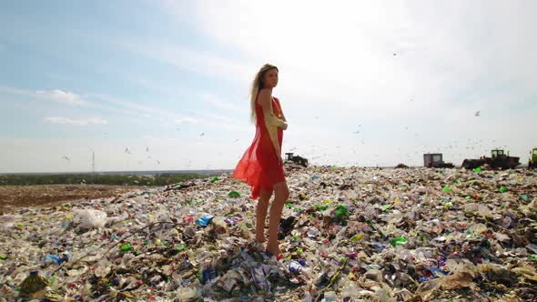 Young Female Pollution Activist in Red Dress Standing at Huge Trash Dump