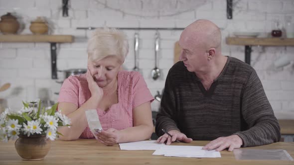 Sad Mature Caucasian Woman Settling Bills with Husband. Senior Man Calming Down His Wife and Smiling