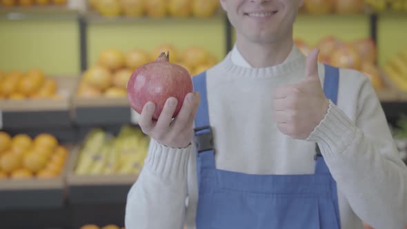 Cheerful Unknown Caucasian Man in Blue Workrobe Holding Pomegranate in Hand and Showing Thumb Up