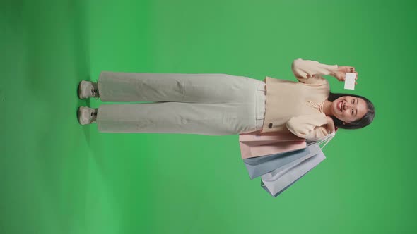 Full View Of Asian Woman Holding A White Card And Shopping Bags Standing In Front Of Green Screen