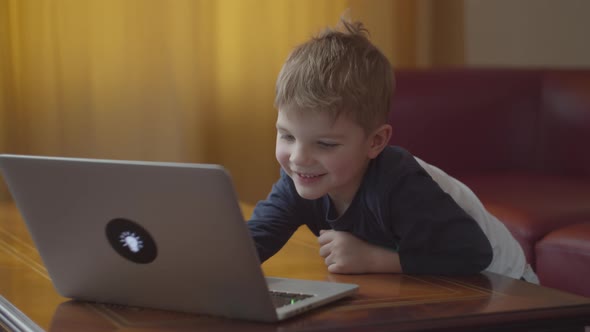 Blonde Preschool Boy Typing on Laptop and Smiling Looking at Computer Screen. Kid Learning Online at