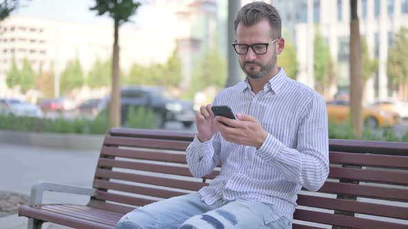 Young Adult Man Using Smartphone While Sitting Outdoor on Bench