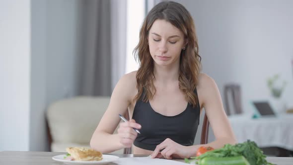 Concentrated Slim Motivated Young Woman Writing Calories Looking at Burger on Plate