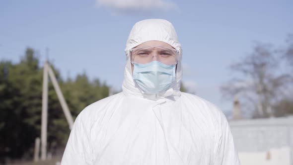 Young Confident Caucasian Man in White Safety Suit Walking in Suburb. Serious Virologist in Face