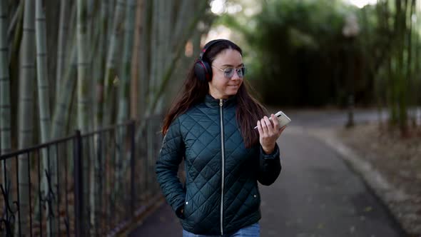 A Smiling Adult Woman with Headphones Listens to Music and Dances Amusingly in a City Park