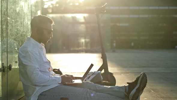 Bearded Middleaged Man in Dark Glasses Sits on the Sidewalk with a Laptop Listening to Music and