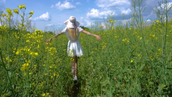 A Young Beautiful Girl in a White Dress in a Wide Hat Walks Through a Flowering Rapeseed Field