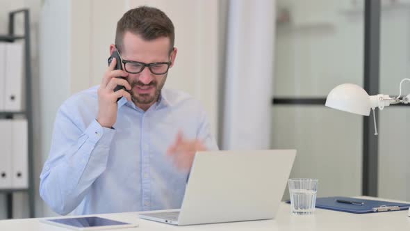 Middle Aged Man Angry on Smartphone While Working on Laptop