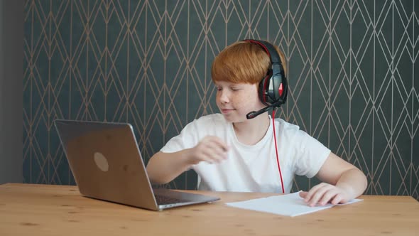 Distance Learning Redhaired Teen Boy Sitting on the Table in Living Room and Uses a Laptop to Study