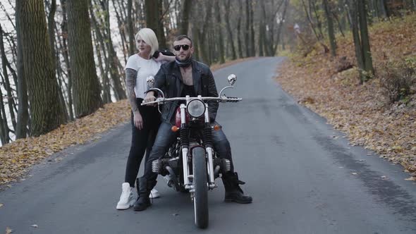 Portrait of Brutal Couple Stands in Fixed Pose at Motorbike and Looks Aside